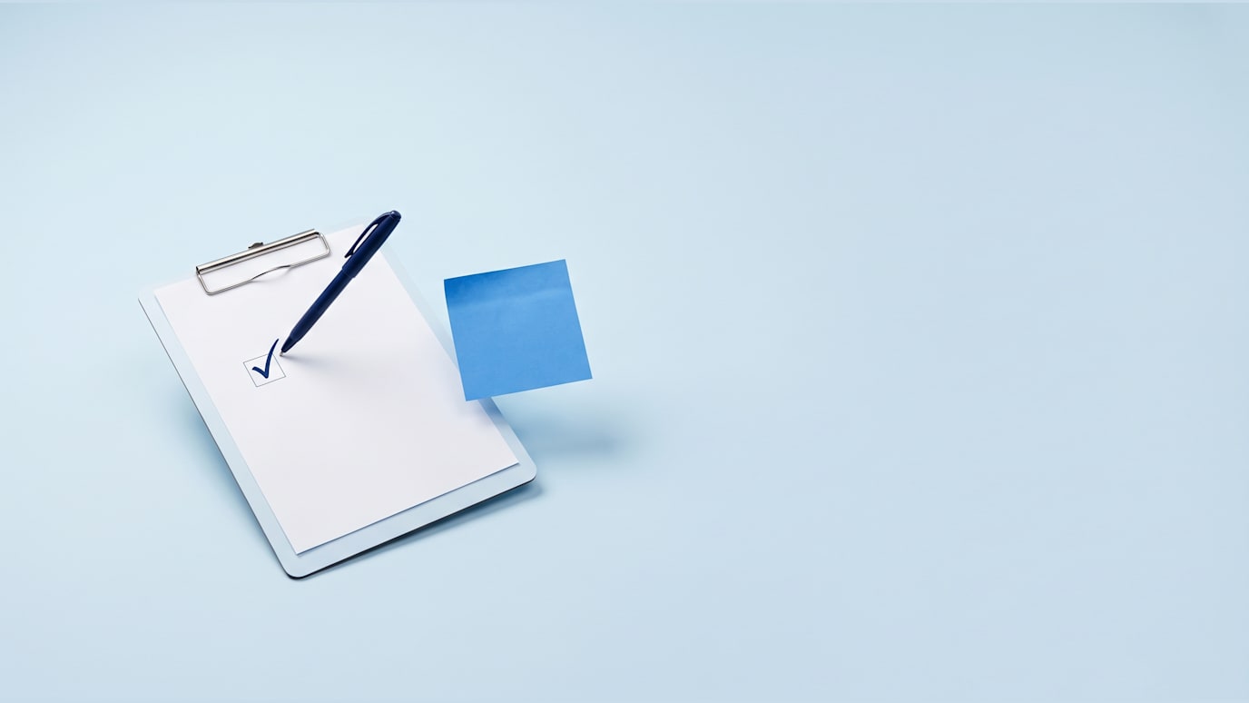 A checklist is being ticked with a navy pen as a blue sticky note is moved to the next task