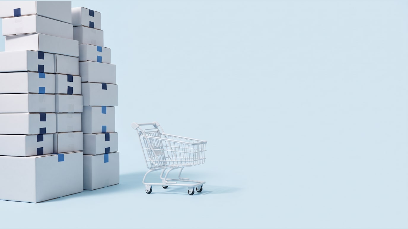 A small white shopping cart rolling away empty from a tall pile of identical sealed white boxes